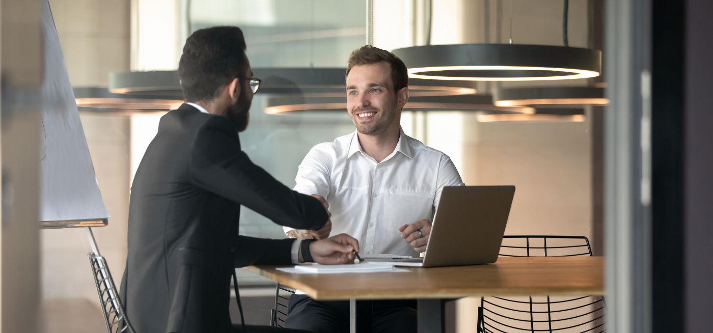 Aperto de mão entre dois profissionais numa sala de reuniões, com portátil sobre a mesa